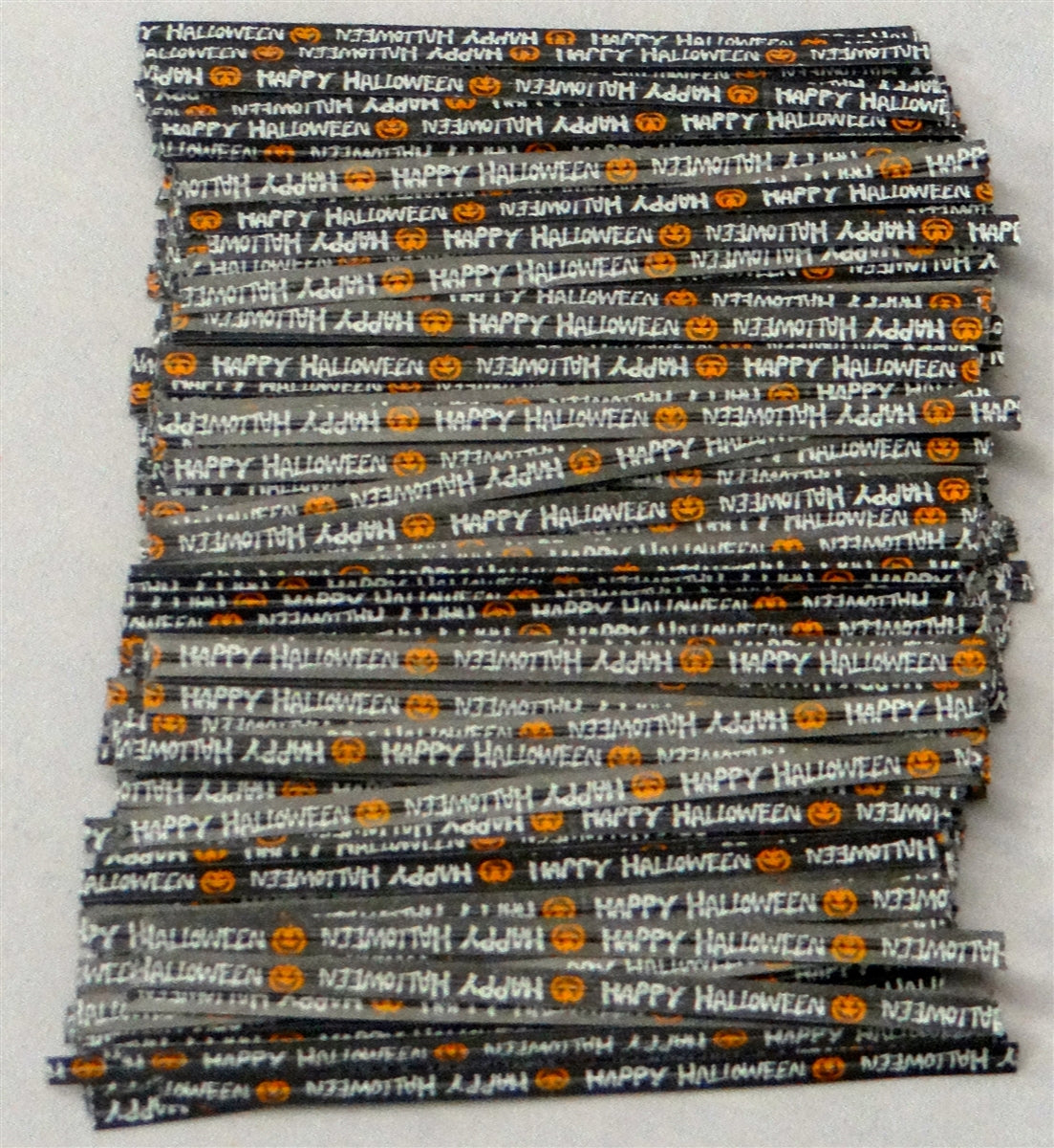 A stack of long paper twist tie with a Halloween theme, featuring the repeated phrase 'Happy Halloween' in alternating orientation, with pumpkin emojis in between. The background is black, with the text and emojis in a contrasting orange, commonly associated with Halloween. This festive twist tie is laid out straight against a light grey background, clearly displaying the celebratory message and design.