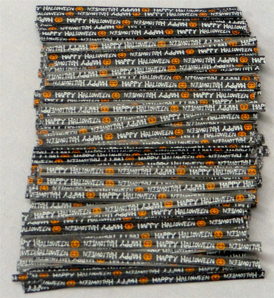 A stack of long paper twist tie with a Halloween theme, featuring the repeated phrase 'Happy Halloween' in alternating orientation, with pumpkin emojis in between. The background is black, with the text and emojis in a contrasting orange, commonly associated with Halloween. This festive twist tie is laid out straight against a light grey background, clearly displaying the celebratory message and design.