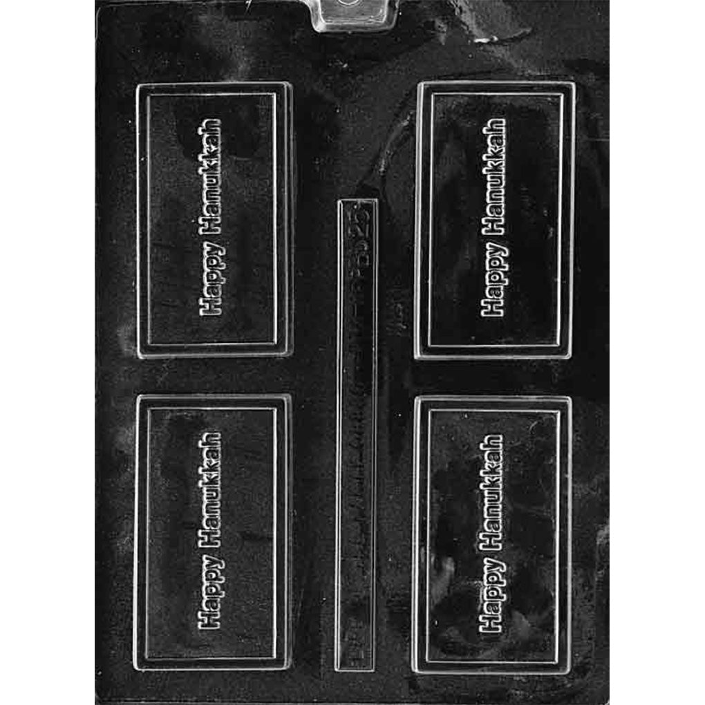 Clear plastic chocolate mold with four rectangular bar cavities embossed with the words “Happy Hanukkah” inside a raised border, plus a narrow rectangular strip cavity in the center, on a dark background.