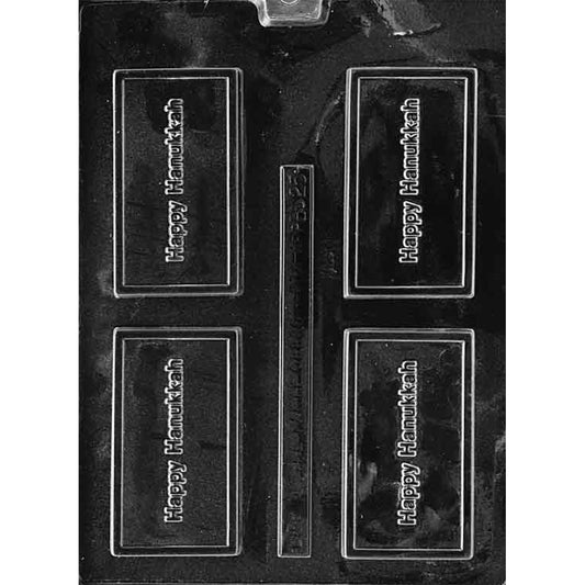 Clear plastic chocolate mold with four rectangular bar cavities embossed with the words “Happy Hanukkah” inside a raised border, plus a narrow rectangular strip cavity in the center, on a dark background.