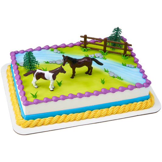 Rectangular cake decorated with two plastic horses, trees, and a fence on a green and blue icing pasture.