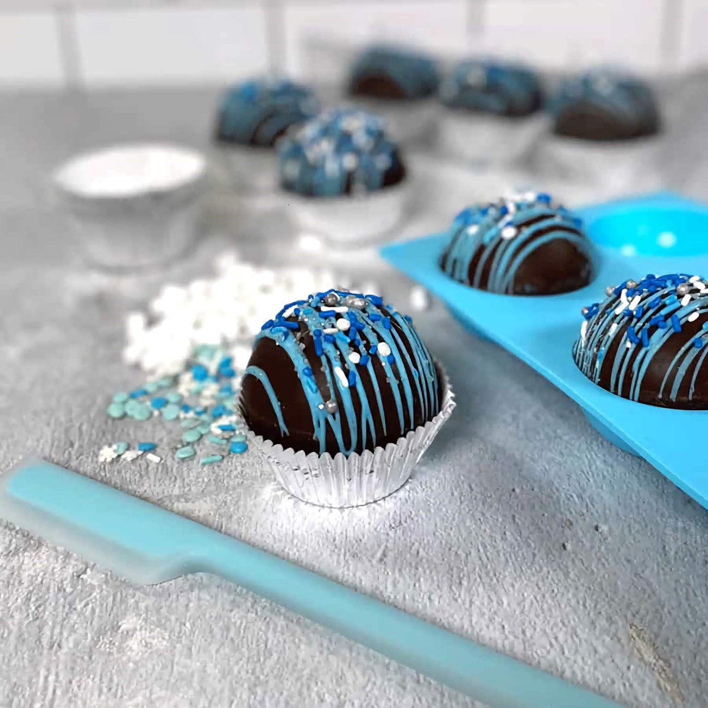 The image presents a culinary scene with completed hot chocolate bombs in the foreground, elegantly drizzled with blue icing and sprinkled with white and blue decorations. These spherical chocolate treats sit in paper cups and are staged on a light-colored countertop. In the background, a light blue silicone mold, the kind used to create these chocolate spheres, can be seen with additional unfinished chocolate bombs in its cavities.