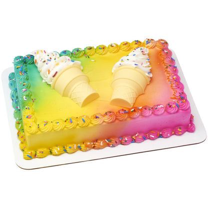 Rectangular cake with rainbow airbrushed icing and piped borders, decorated with two sideways ice cream cone toppers and swirls of white frosting with sprinkles.