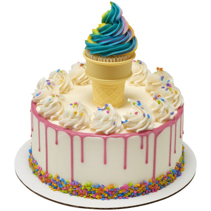 Round white cake with pink drip icing and rainbow sprinkle border, topped with white frosting swirls and a cupcake set inside an assembled yellow ice cream cone topper.