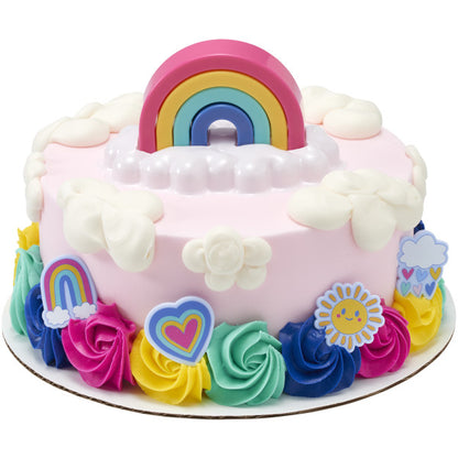 Round pink frosted cake with white cloud dollops and colorful buttercream rosettes around the base, topped with a stacked rainbow on a white cloud tray.