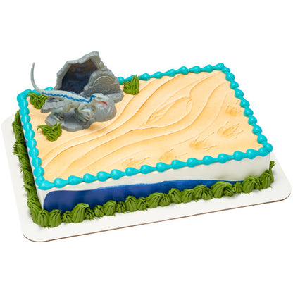 A rectangular cake decorated with sandy textures, dinosaur footprints, and a Blue Raptor cake topper in front of a Jurassic World™ rock formation.