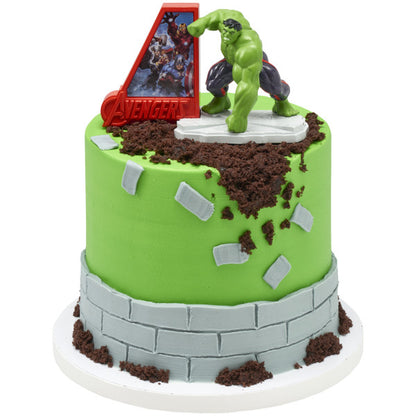 Round green cake styled like cracked concrete with gray brick banding and chocolate crumb rubble, topped with Hulk figure on base and a red Avengers sign.