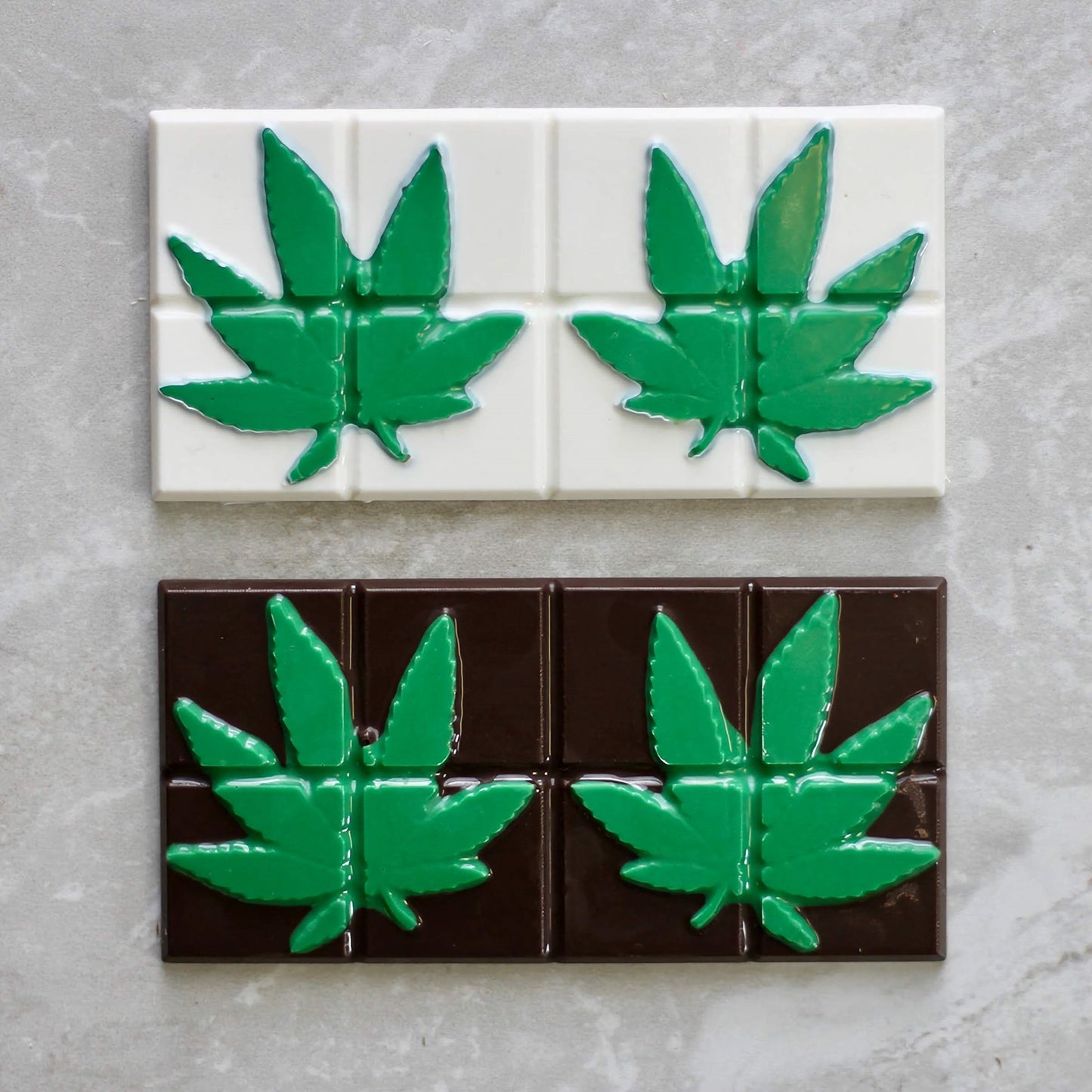 a chocolate mold with segmented squares, each featuring a raised, detailed imprint of a marijuana leaf. The top row of squares is white chocolate with green leaves, and the bottom row is dark chocolate with green leaves, all set against a gray stone background.