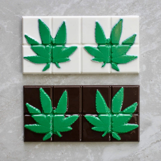 a chocolate mold with segmented squares, each featuring a raised, detailed imprint of a marijuana leaf. The top row of squares is white chocolate with green leaves, and the bottom row is dark chocolate with green leaves, all set against a gray stone background.