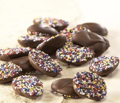 Milk chocolate non-pareils covered with colorful sugar sprinkles, displayed on a rustic surface.