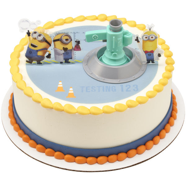 A round cake with a Minions-themed edible image, bordered in yellow and blue frosting, topped with the Minions Fart Bubble Blaster DecoSet.