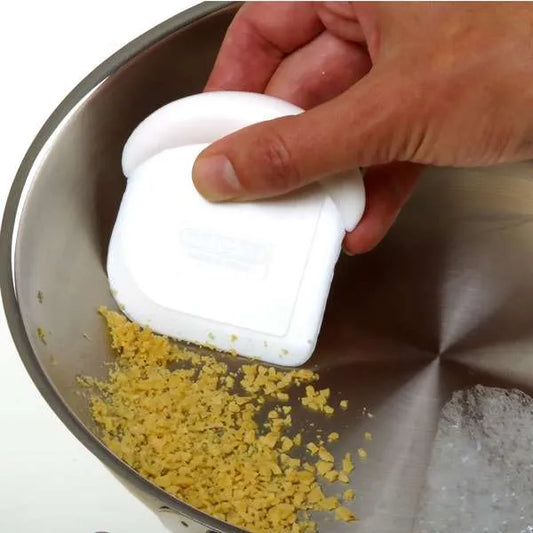 Close-up of a hand using a white plastic scraper to clean food residue from a stainless steel mixing bowl.