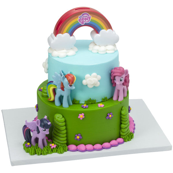 A tiered cake decorated with the My Little Pony™ Signature DecoSet®, featuring a whimsical landscape with the rainbow topper and pony figurines placed around the cake.