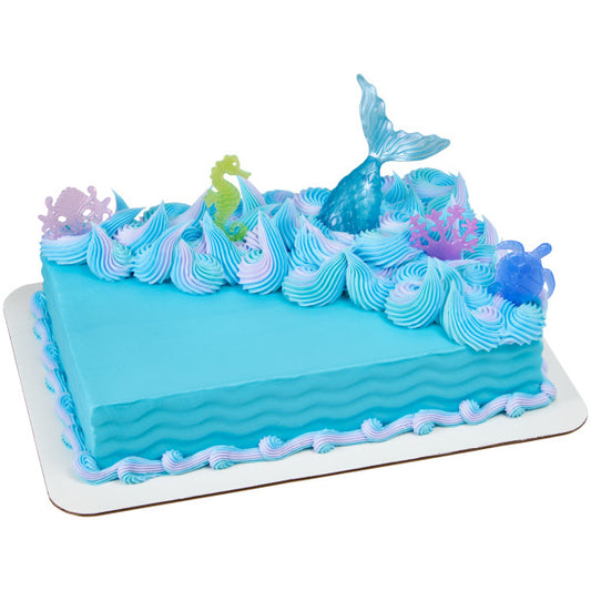 Rectangular blue sheet cake decorated with ocean wave frosting and topped with Mystical Mermaid plastic toppers including mermaid tail, seahorse, turtle, coral, and shell crown.