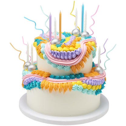 Two-tier white cake decorated with colorful piped buttercream in teal, orange, pink, and purple, topped with pastel twist shaped candles, silver disco ball decorations, and white striped candles.