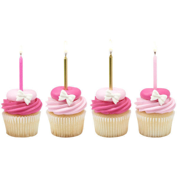 Four cupcakes topped with pink buttercream swirls, heart-shaped decorations with white bows, and tall birthday candles in bright pink, light pink, and metallic gold.