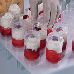 Tray of clear plastic push-up pop dessert cups filled with red fruit layers and white whipped cream, topped with cherries and berries; a gloved hand places a lid on one cup.