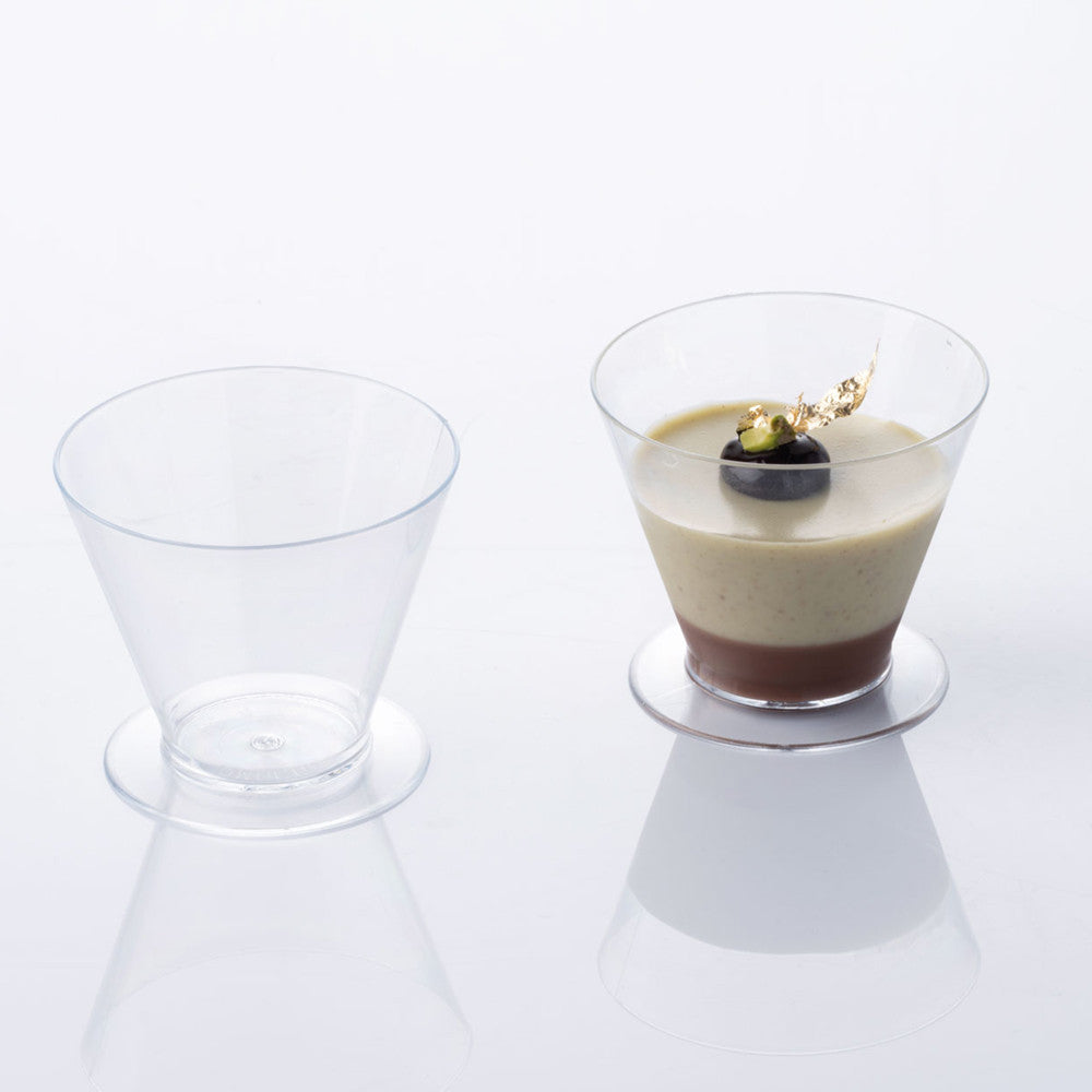 Two clear cone-shaped dessert cups with footed bases on a white reflective surface, one empty and one filled with layered cream and chocolate dessert topped with a blueberry and gold leaf.