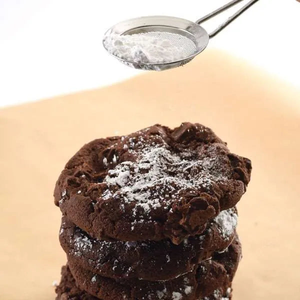 Stainless steel fine mesh strainer sifting powdered sugar over a stack of chocolate cookies on parchment paper.