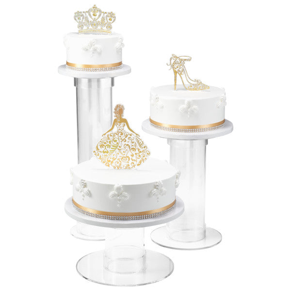 Three elegant white cakes on pedestals, each topped with a different gold Quinceañera-themed topper: dress silhouette, crown, and shoe.