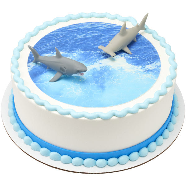 A round cake with a realistic water-themed edible image, bordered in blue frosting, and topped with the Shark Attack DecoSet shark figurines.