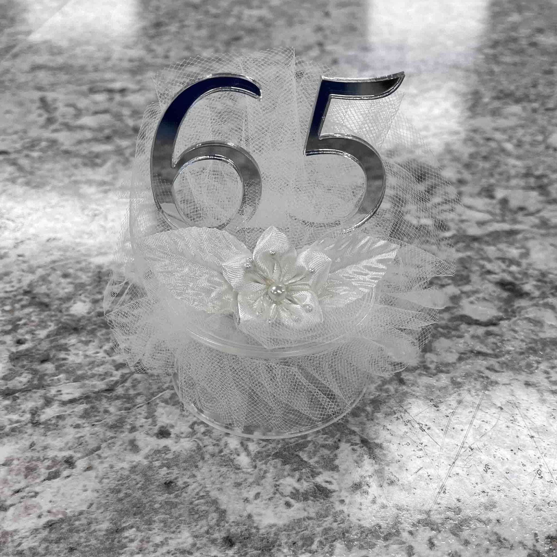 Elegant silver '65' numeral cake topper for a 65th-anniversary celebration, featuring a delicate white floral embellishment and tulle detail, presented on a clear base, perfect for a commemorative cake centerpiece.