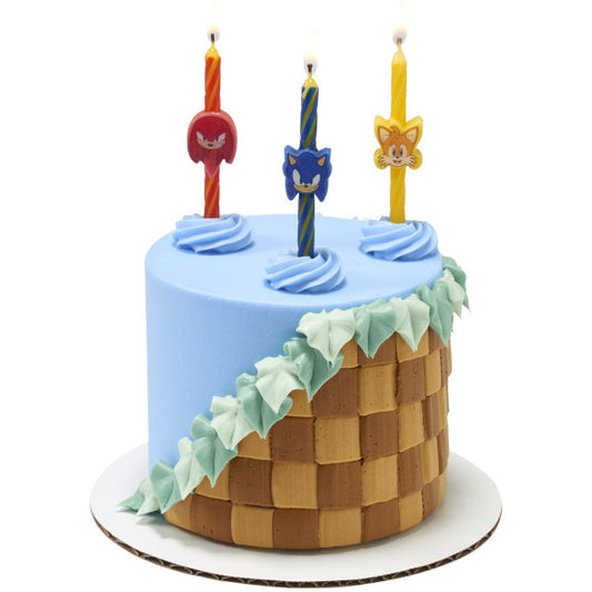 Light blue frosted round cake with a brown checkerboard side design and green piped border, topped with three lit Sonic character candles for Knuckles, Sonic, and Tails.