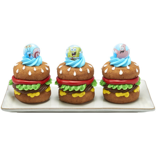 Three burger-shaped cupcakes on a rectangular plate, each topped with a bright blue frosting swirl and a SpongeBob SquarePants themed ring topper in a round blue design.