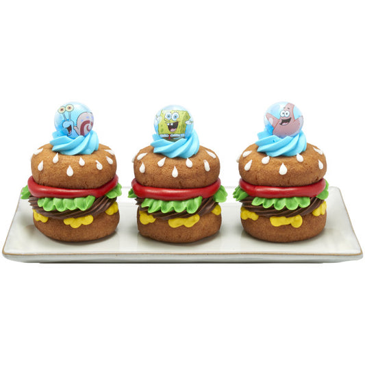 Three burger-shaped cupcakes on a rectangular plate, each topped with a bright blue frosting swirl and a SpongeBob SquarePants themed ring topper in a round blue design.
