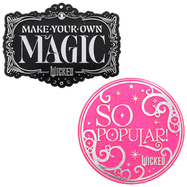 Two decorative Wicked cake plaques on a white background: a black ornate rectangle reading “MAKE YOUR OWN MAGIC” in metallic silver and a bright pink circular plaque reading “SO POPULAR,” both with swirly star accents.