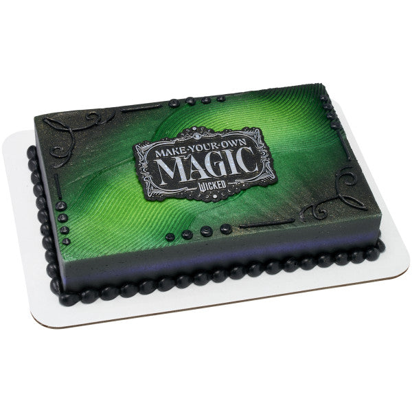 Rectangular green sheet cake with black piped border and scroll accents, topped at center with the silver-on-black “MAKE YOUR OWN MAGIC” Wicked plaque.