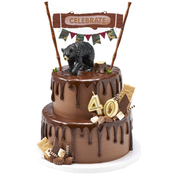 A camping-themed cake with a black bear topper, a "Celebrate" banner, and s'mores decorations.