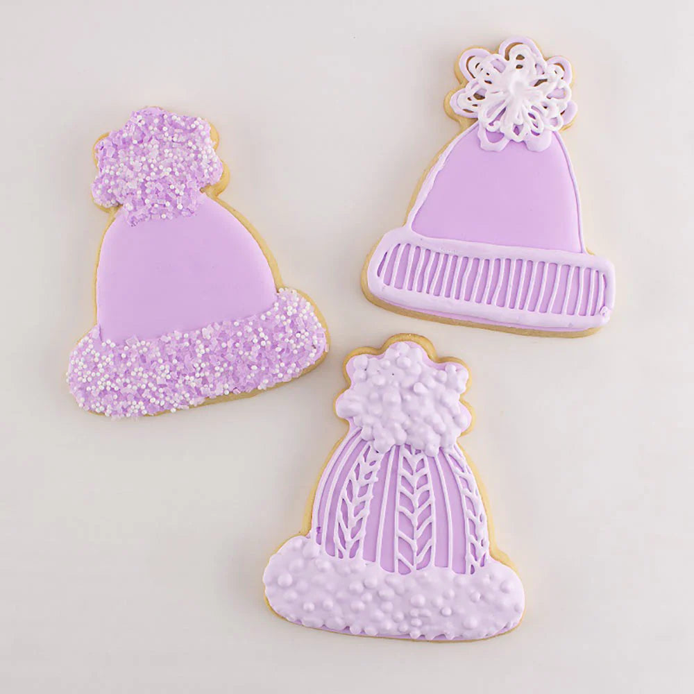 Three decorated winter hat cookies in lavender and white icing with piped details and sprinkles, shaped using the Ann Clark cookie cutter