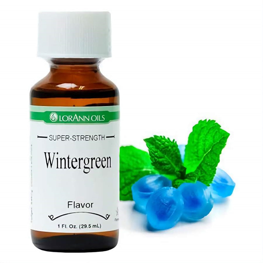 Amber bottle of LorAnn Super Strength Wintergreen Flavor, accompanied by vibrant green mint leaves and blue wintergreen candies, ideal for adding a fresh, minty taste to candies and baked treats.