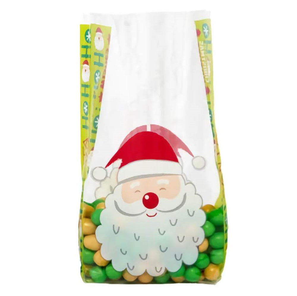 A large cellophane treat bag with a 'Wishes For Santa' theme, filled partway with green and gold candies. The bag features a jolly Santa face with a full white beard and red hat at the center, flanked by colorful Christmas motifs and greetings on the sides. The transparent portion above Santa's face allows the contents to be seen, enhancing the festive design suitable for holiday gifting.
