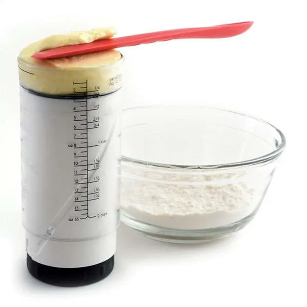 Adjustable measuring cup filled with a thick ingredient, shown with the plunger pushed upward beside a mixing bowl and red spatula.