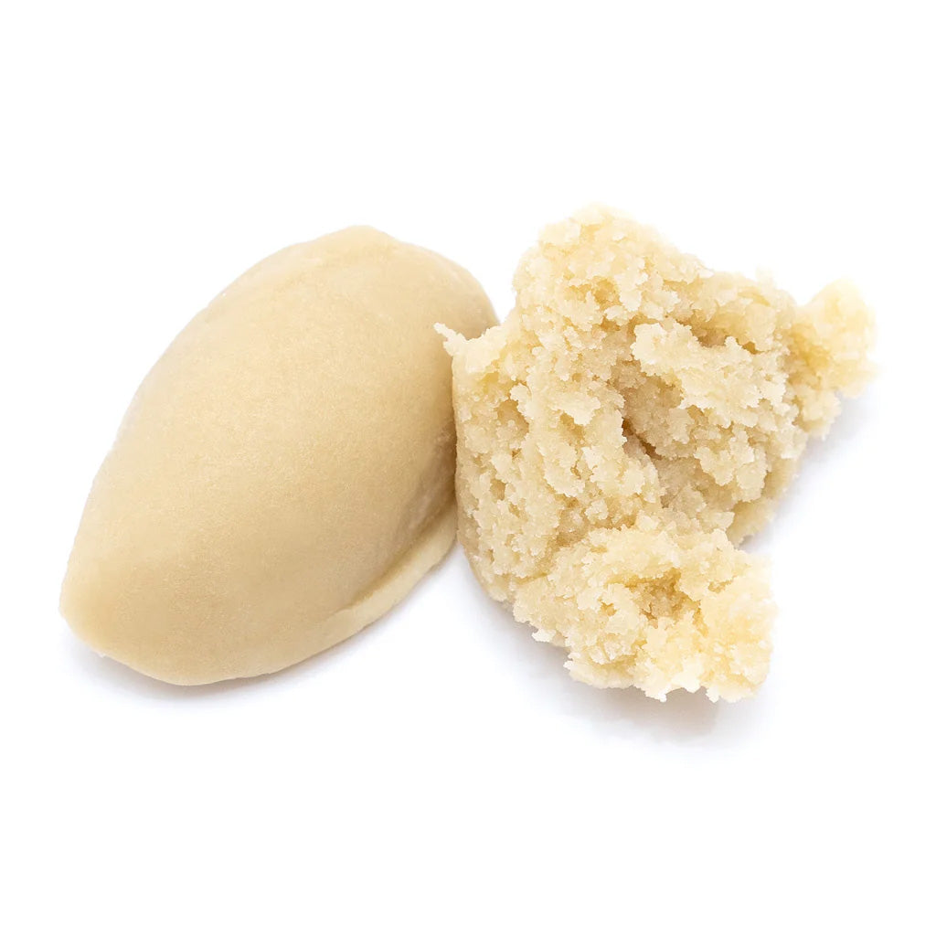 Beige almond paste shown on a white background, with one smooth oval piece and one crumbly broken piece revealing a soft, grainy interior.
