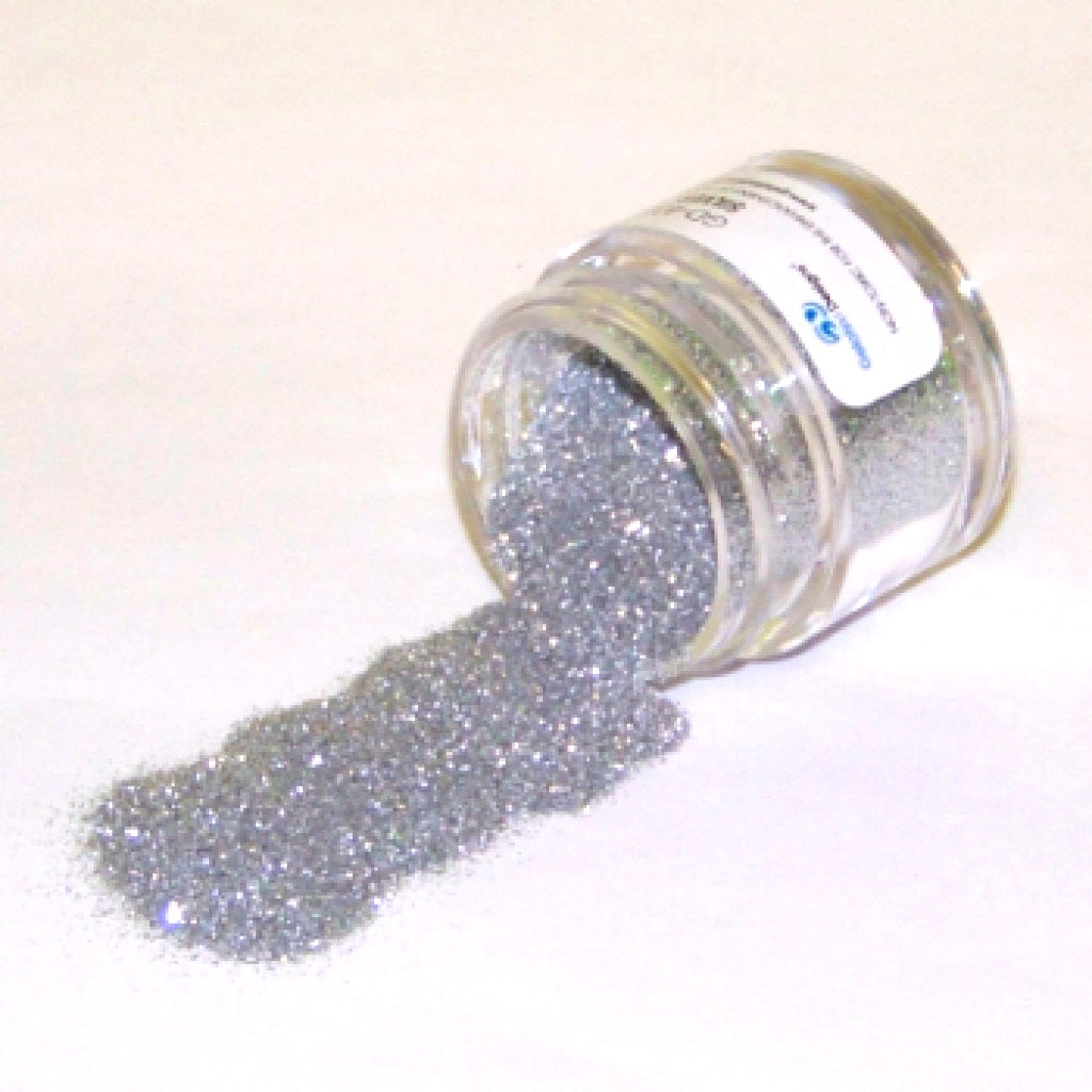 Small clear jar tipped on its side spilling ultra-fine silver glitter dust onto a white surface, with a soft metallic sparkle and a partially visible label.