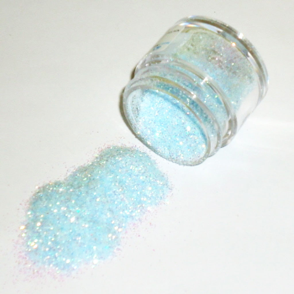 Small clear plastic jar tipped on its side with baby blue iridescent glitter dust spilling onto a white surface, showing fine sparkly particles with rainbow reflections.