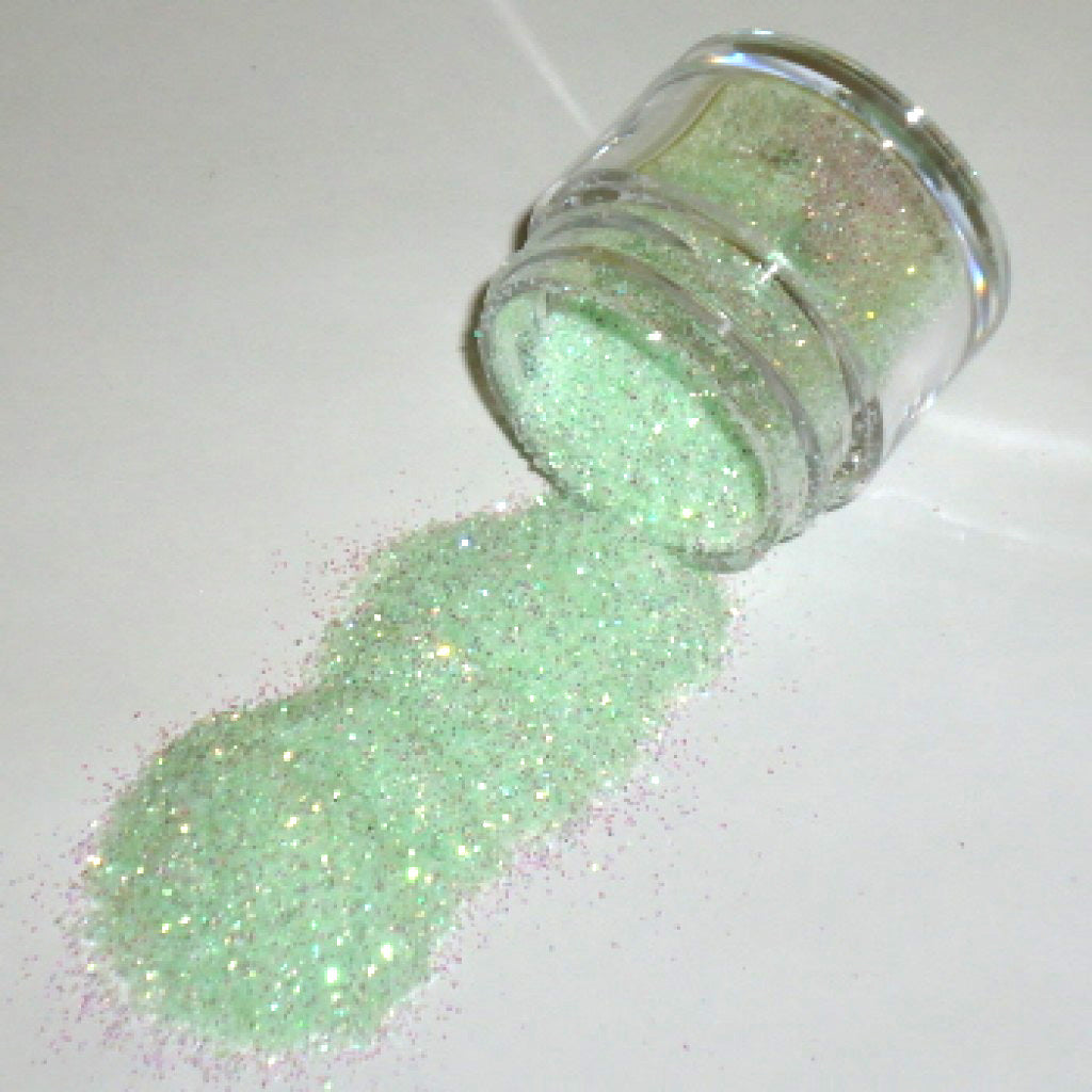 Small clear jar tipped on its side spilling baby green glitter dust onto a white surface, showing fine pale green sparkling particles with soft iridescent highlights.