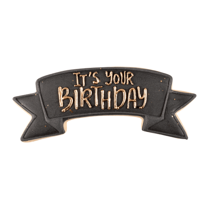 Banner-shaped cookie decorated in black and gold with 'It’s Your Birthday' written in icing.