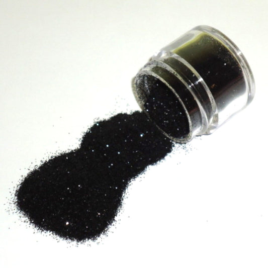 Small clear jar tipped on its side spilling black glitter dust onto a white surface, with fine sparkling particles visible throughout.