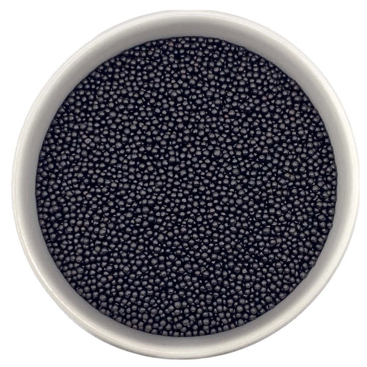 Top-down view of a white bowl filled with black non-pareils, showing tiny round sprinkle beads in a deep charcoal-black shade.