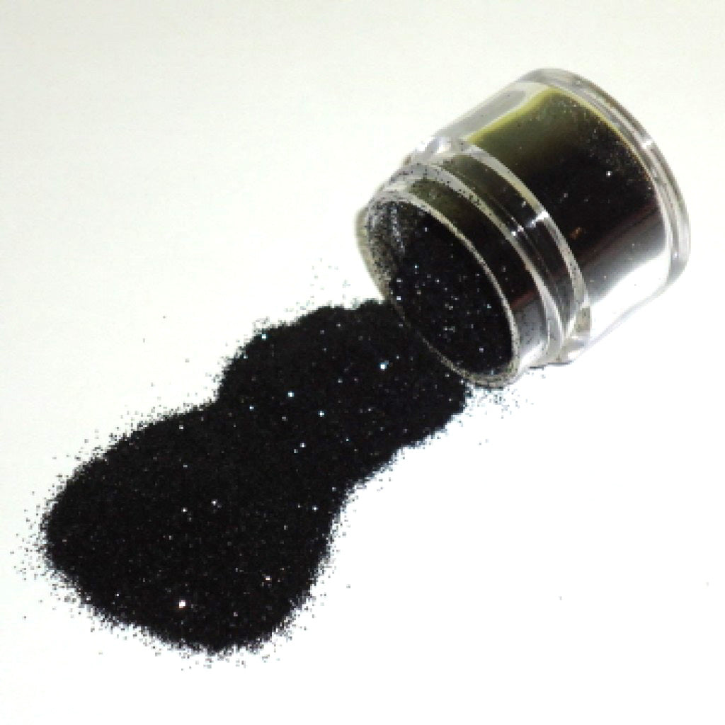 Small clear jar tipped on its side spilling black sparkle glitter dust onto a white surface, with fine shimmering particles visible throughout.