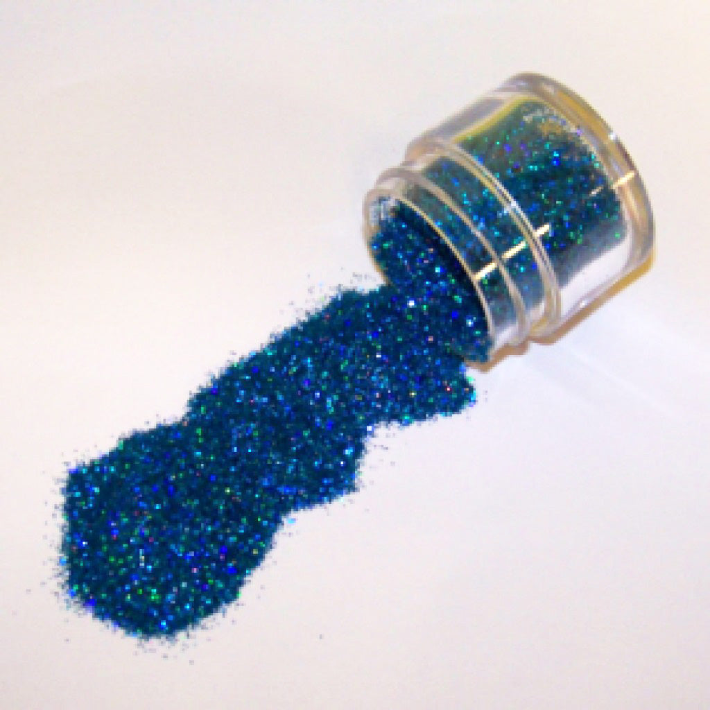 Small clear jar tipped on its side spilling blue hologram glitter dust onto a white surface, with fine sparkling particles reflecting blue and rainbow highlights.