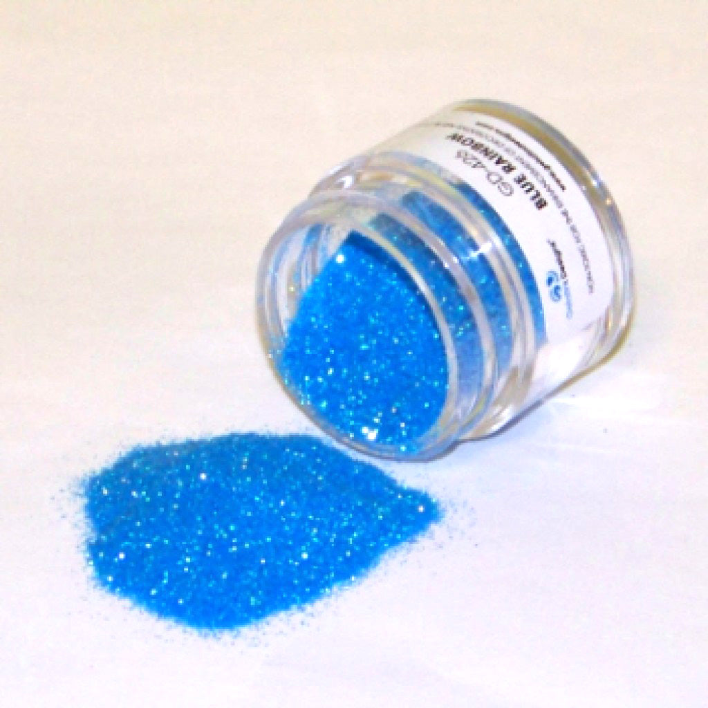 Small clear jar tipped on its side spilling bright blue glitter dust onto a white surface, showing fine sparkling blue particles with rainbow shimmer.