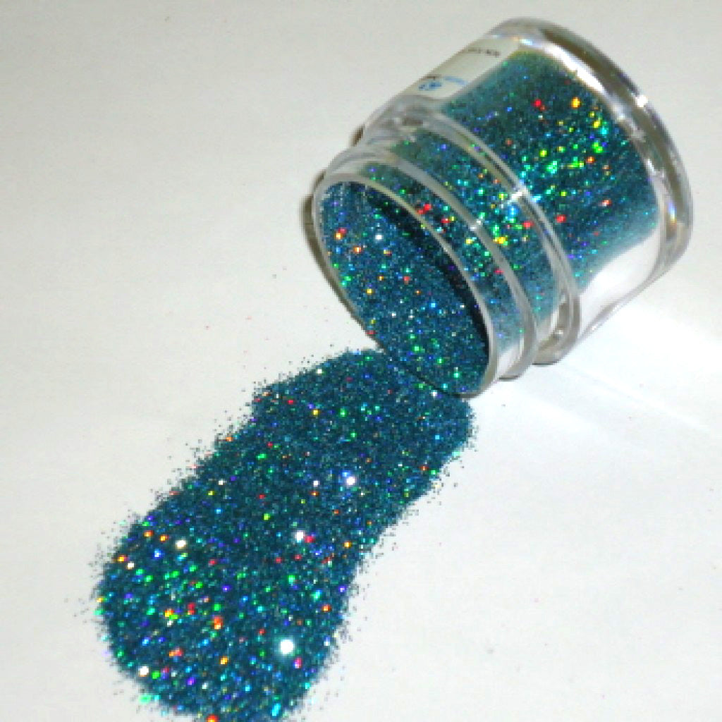 Blue Topaz Galaxy Glitter Dust spilled from a small clear jar, showing sparkling blue glitter with rainbow reflectivity on a white background.