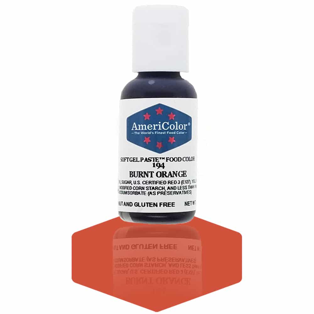 A bottle of AmeriColor Burnt Orange soft gel paste food coloring with a white cap and blue label featuring red stars, standing on a deep burnt orange reflective surface that mirrors the bottle.