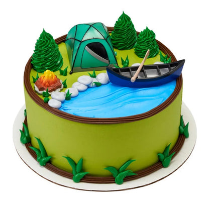 Campfire, tent, and canoe cake kit arranged on a round camping cake with blue icing water, piped evergreen trees, grass, and rocks
