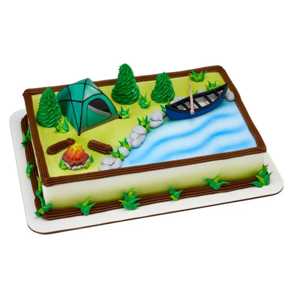 Camping cake kit on a sheet cake decorated with a blue icing river, piped evergreen trees, rocks, logs, and green grass